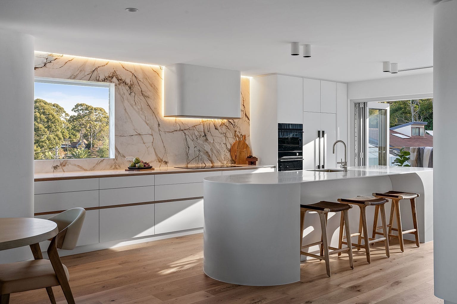 Modern luxury kitchen with a curved white island, marble backsplash with backlighting, integrated appliances, timber stools, and large windows bringing in natural light.